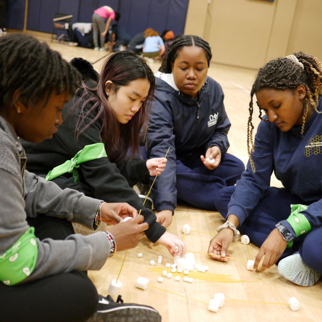 Four young women sitting on floor in gym building a pasta and marshmallow tower