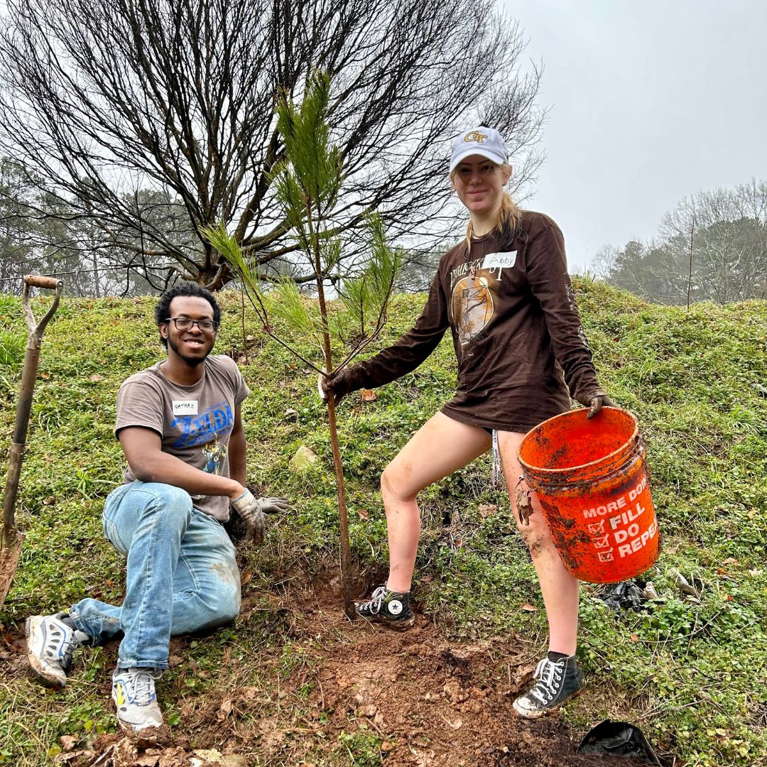 one young man and one young woman standing and smiling next to a freshly planted tree