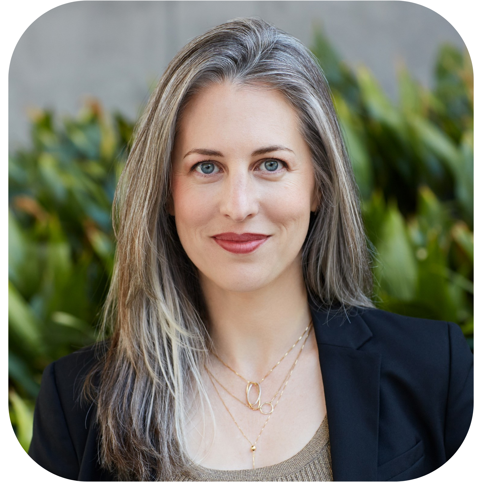 Brynn Barineau is a white woman with long, straight gray hair. She's wearing a dark blazer over a dark beige shirt and she's standing in front of vibrant green bushes.