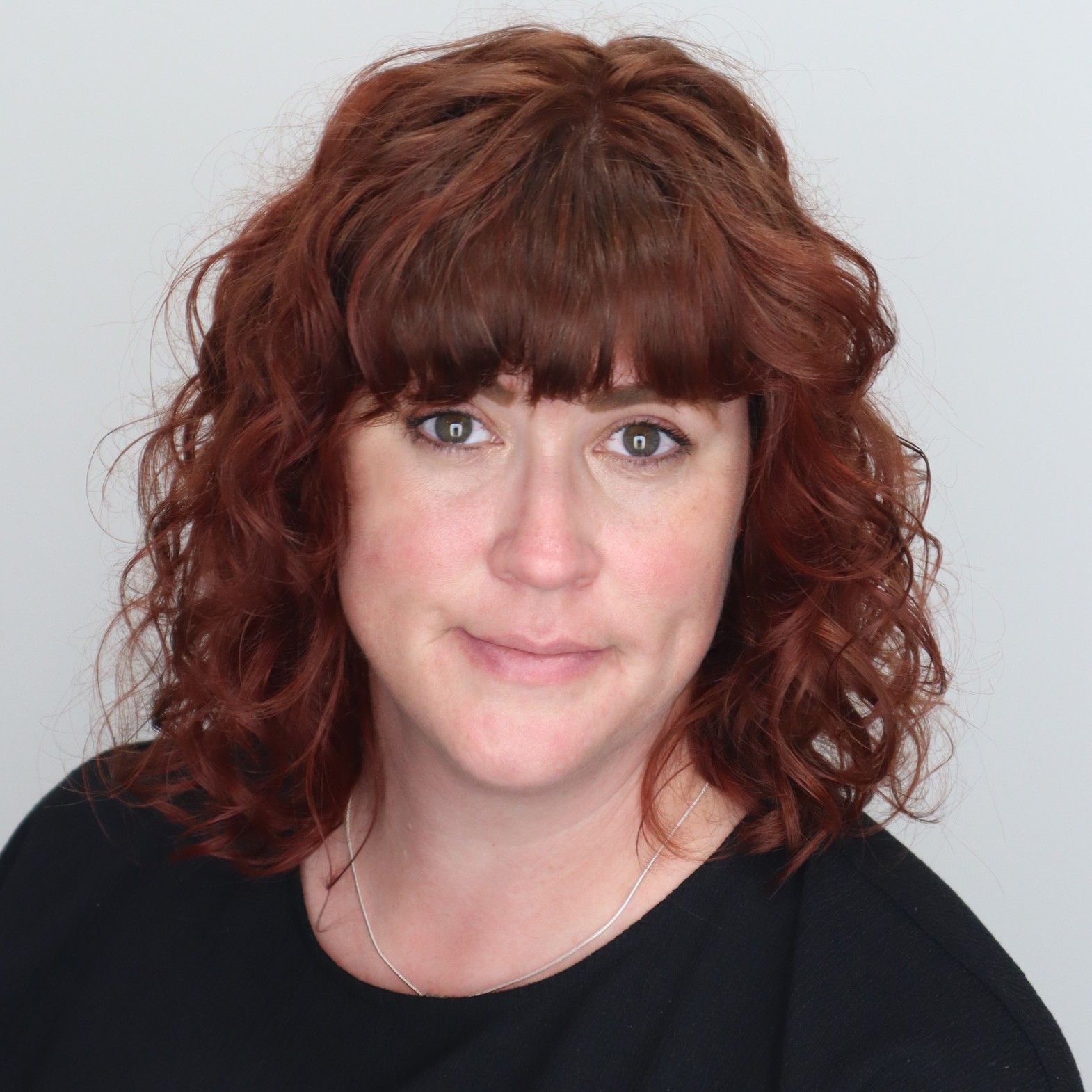 Ashlee Flinn is a white lady with red curly hair and bangs who is smirking at the camera with a closed lip smile