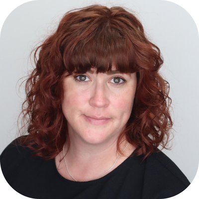 Ashlee Flinn is a white lady with red curly hair and bangs who is smirking at the camera with a closed lip smile